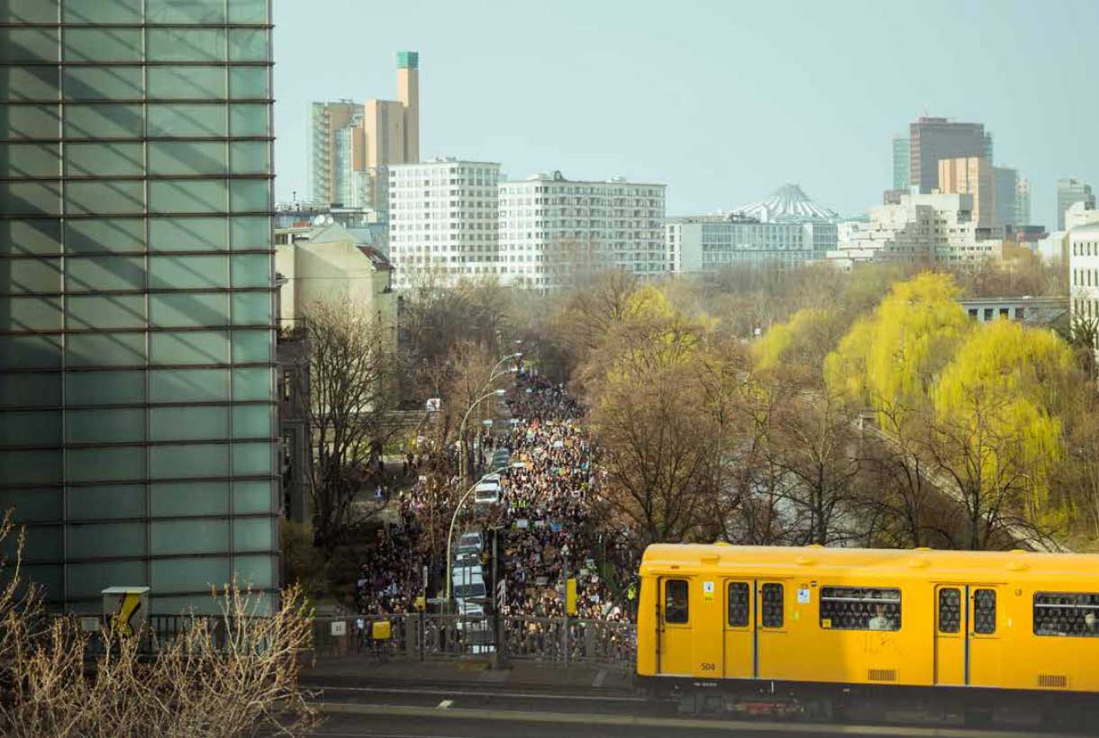 Eine für Berlin typische gelbe U-Bahn fährt an einer belebten Straße mit Hochhäusern im HIntergrund